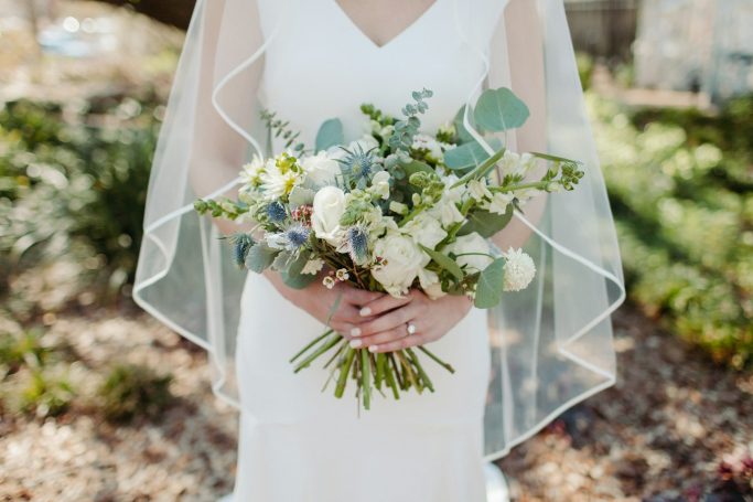 Braut mit weißem Kleid und transparentem Schleier, hält einen Blumenstrauß.