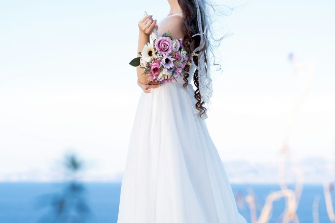 Braut in langem weißen Kleid mit Blumenstrauß vor blauem Wasserhintergrund.