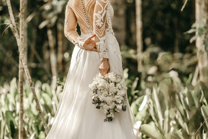 Braut in eleganter, weißer Hochzeitkleid hält einen Blumenstrauß, umgeben von Natur.