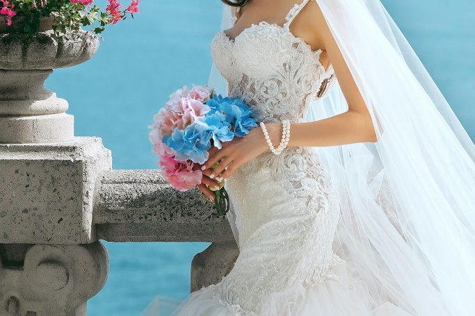 Braut in einem eleganten Kleid mit buntem Blumenstrauß vor blauem Wasser.
