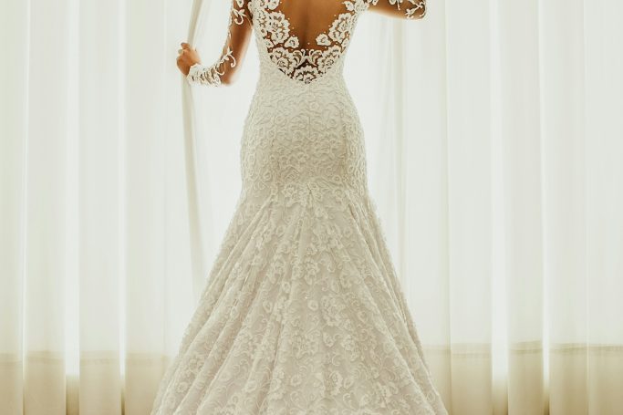 Braut in einem eleganten, romantischen Kleid mit Spitzenrücken vor einem hellen Vorhang.