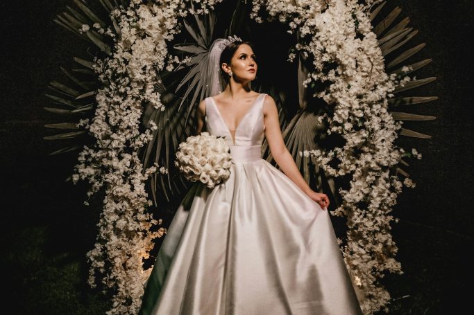 Braut in eleganter, schneeweißer Robe vor einer Wand aus weißen Blumen.