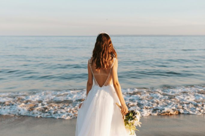 Braut in einem weißen Kleid steht am Strand und schaut auf das ruhige Meer.