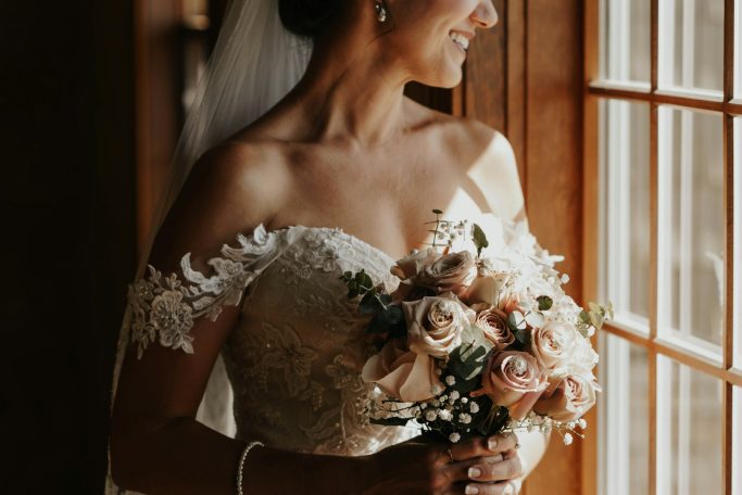 Braut mit Bouquet in der Hand, steht vor einem Fenster und blickt nachdenklich hinaus.